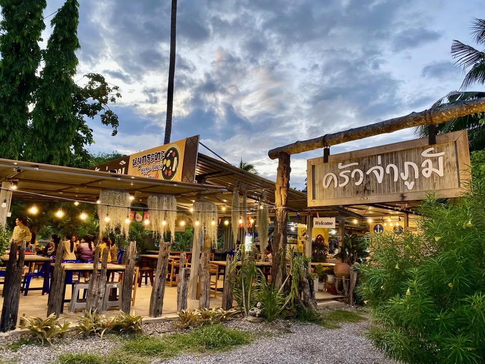 ป้ายไม้หน้าร้านและการตกแต่งไฟวอร์มไลท์ยามเย็นของร้านครัวจ่าภูมิ หมูกระทะและอาหารตามสั่งสไตล์บ้านๆ