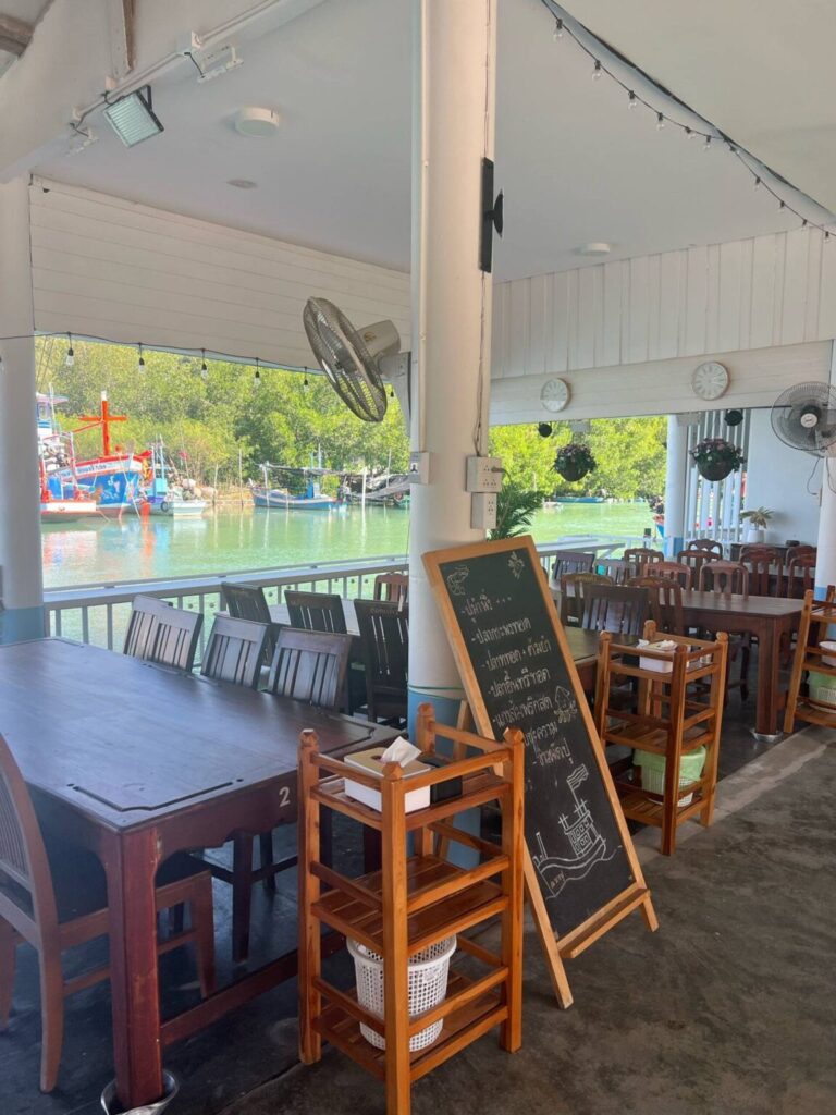 บรรยากาศทางเดินสะพานไม้ริมคลองและเรือประมงของร้านอาหาร ณ บางปู สามร้อยยอด เบื้องหลังเป็นวิวภูเขาและหมู่บ้านชาวเล